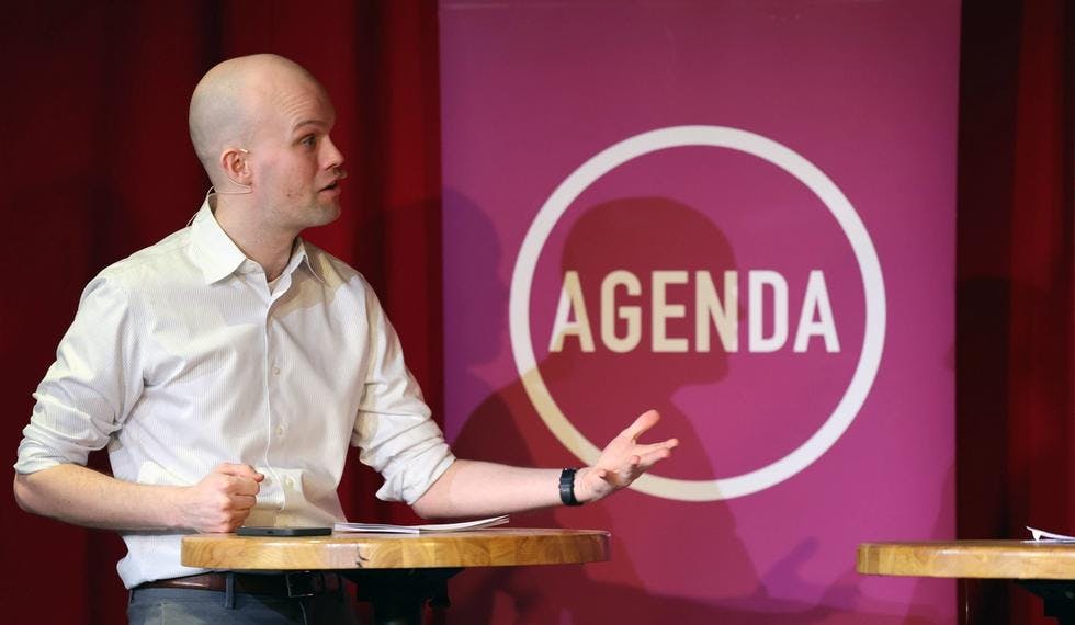 Bilde av Axel Fjeldavli ved et høyt ståbord. Han gestikulerer og ser ut som han er midt i å si noe. Bak han står en rollup med Agenda-logoen i lilla og hvitt. 