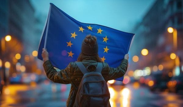 Jente går på gata mens hun holder et stort EU-flagg. Det er blått med gule stjerner og blafrer i vinden. 