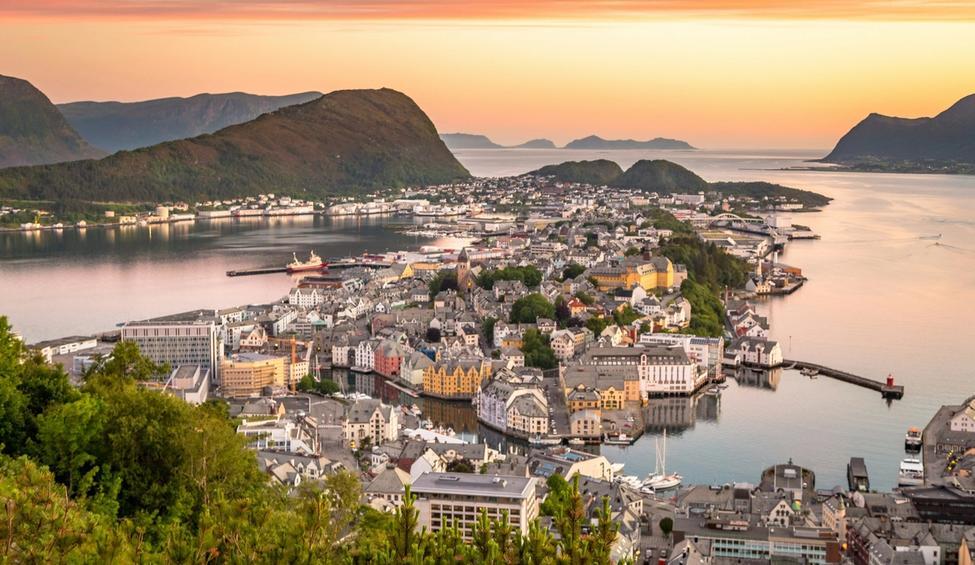 Et bilde av Ålesund, fjord, fjell og by. Det er blankt vann og oransje himmel.